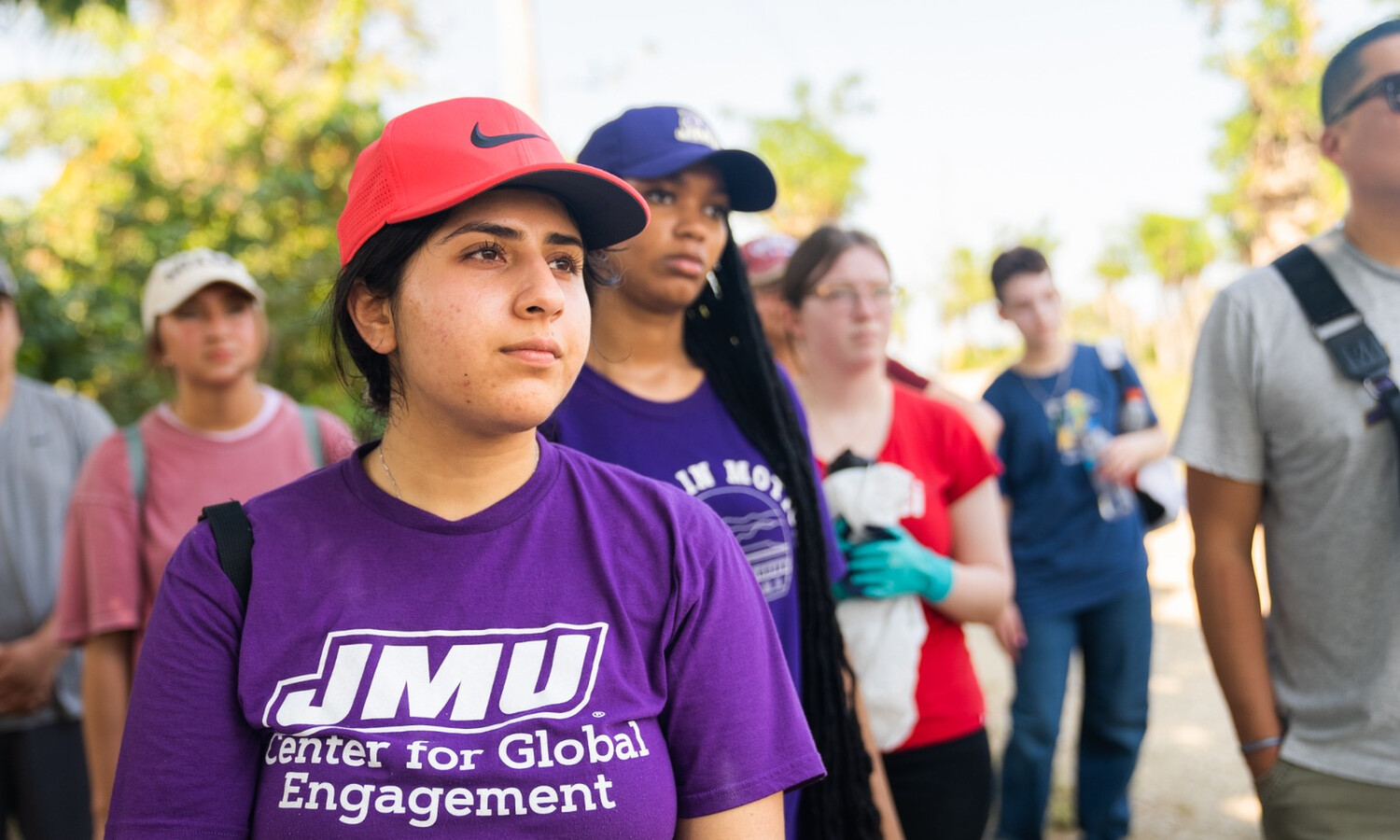 Shukrana Shukrulla wearing red Nike baseball cap and purple JMU T-shirt staring into distance. Other students stand behind her in the background.